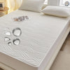 A white fitted mattress protector with a subtle wavy texture is displayed on a bed, next to a pair of headphones.