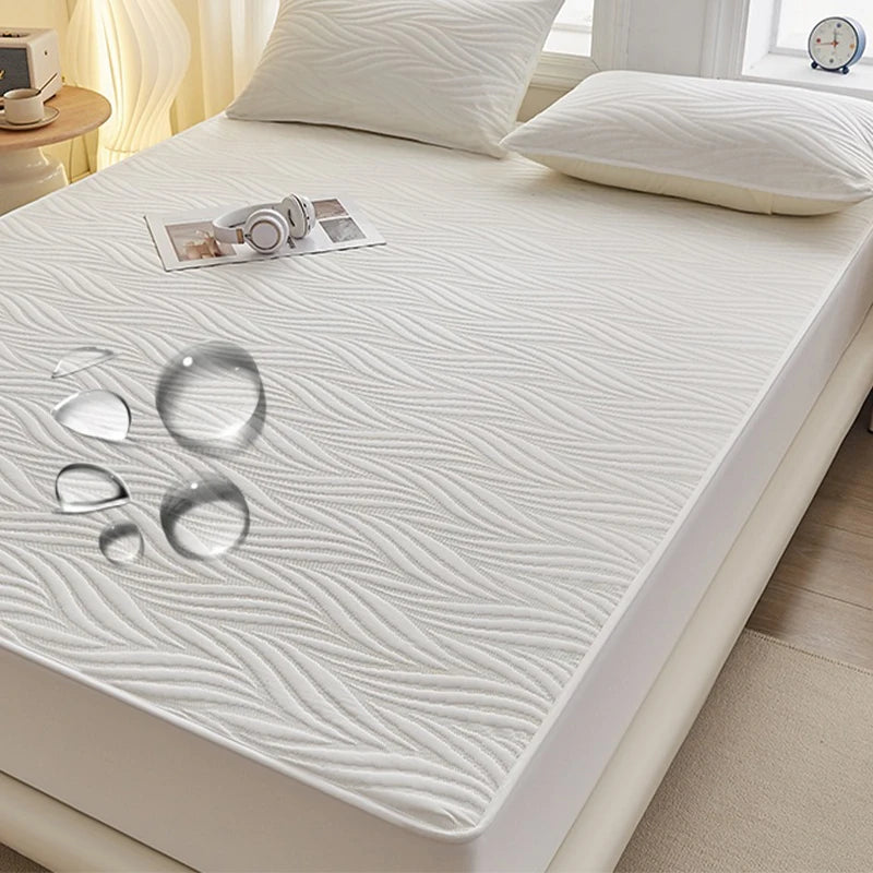 A white fitted mattress protector with a subtle wavy texture is displayed on a bed, next to a pair of headphones.