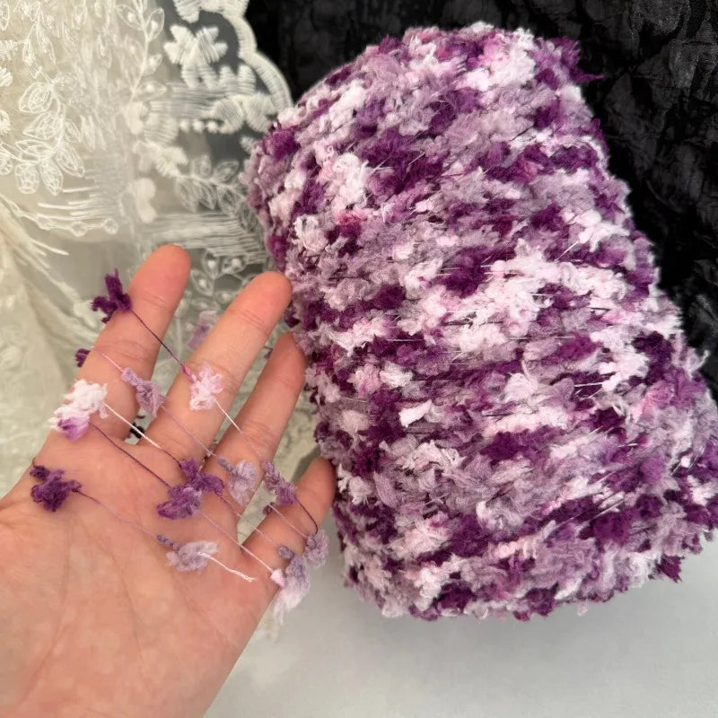 A hand holds a skein of purple and white variegated cotton blend yarn for knitting and crochet.