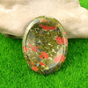 A smooth, oval natural crystal stone with green and red inclusions rests on bright green grass next to a hand.