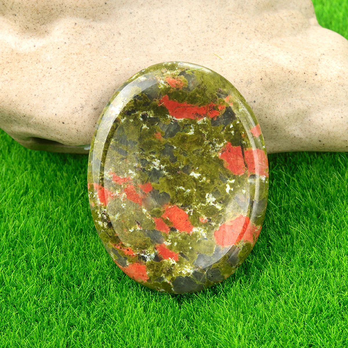 A smooth, oval natural crystal stone with green and red inclusions rests on bright green grass next to a hand.
