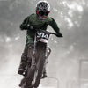 Fahrer mit taktischen Motorradbrillen und grünem Equipment springt auf ein Dirtbike in nebligem Freien.