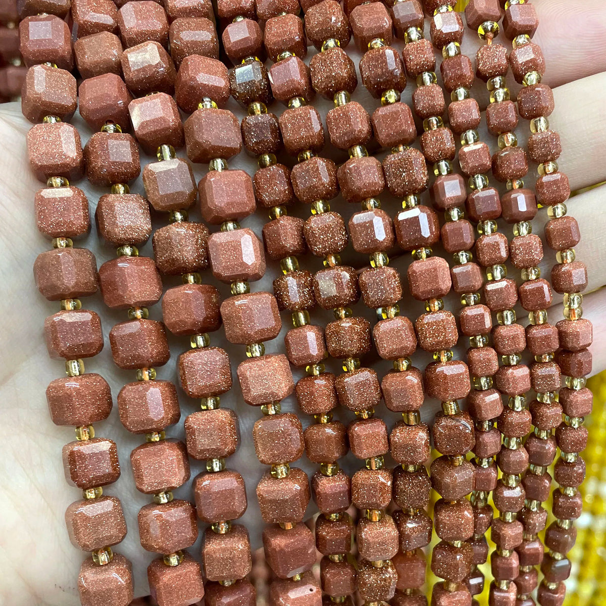 A hand holds a strand of reddish-brown natural stone square faceted beads for jewelry making.