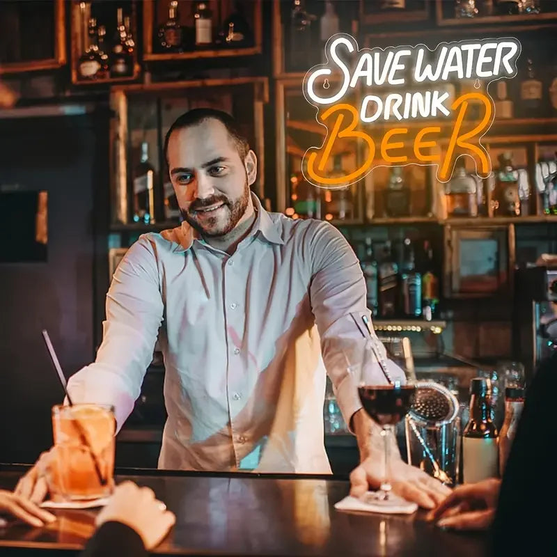 A man in a shirt stands in a bar setting with an LED neon sign that reads Save Water Drink Beer