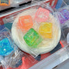 Ein durchsichtbarer Plastikbehälter mit mehreren farbigen quadratischen Stressball-Spielzeugen.
