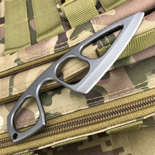 A silver compact fixed-blade knife with a serrated blade and a handle is shown against a camouflage fabric background.