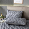 A gray quilted pillow protector with a diamond pattern resting on a bed, next to a headboard and a small green vase.