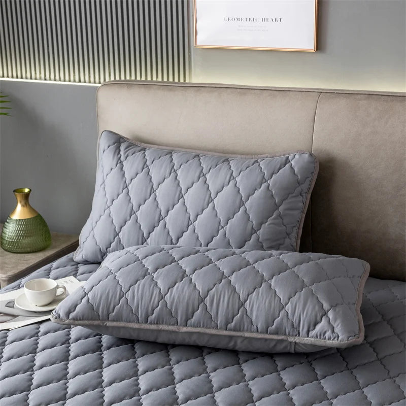 A gray quilted pillow protector with a diamond pattern resting on a bed, next to a headboard and a small green vase.