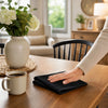 A hand places a black microfiber cleaning towel on a wooden table next to a mug and flowers.