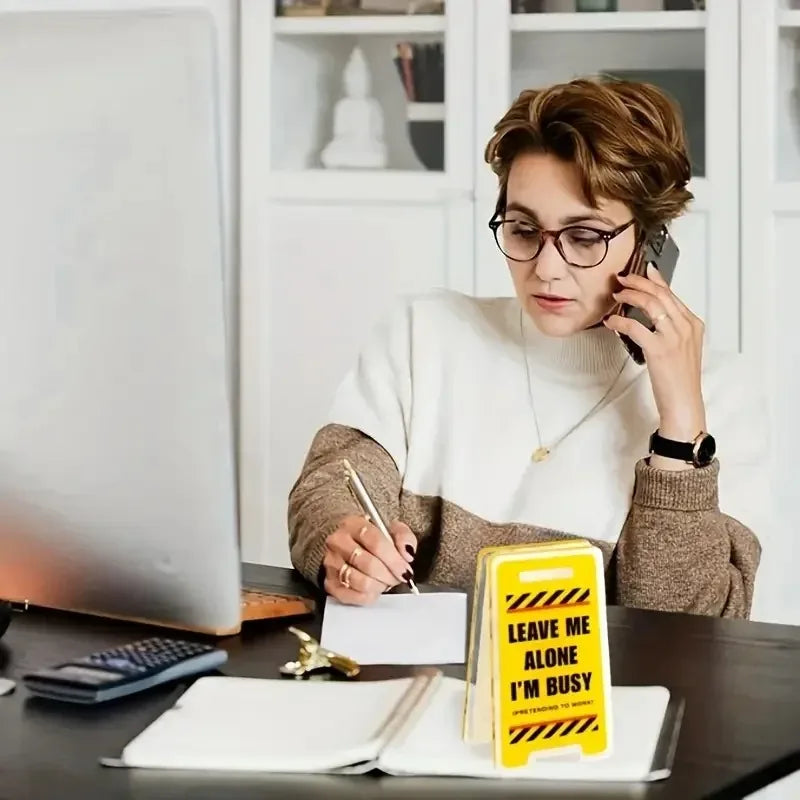 A woman in glasses is writing on a notepad while holding a phone, next to an acrylic desk sign that reads Leave Me Alone I'm 