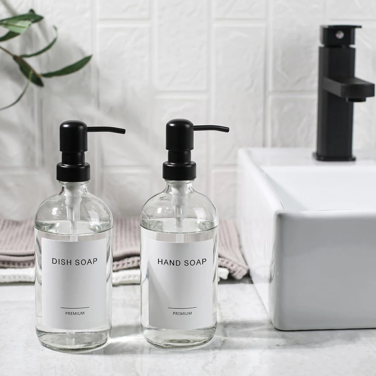 A set of two clear glass soap dispensers with black pumps for dish soap and hand soap on a white countertop.