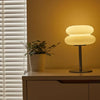 A modern cloud-shaped bedside table lamp with a light shade sits on a wooden surface next to a window blind.