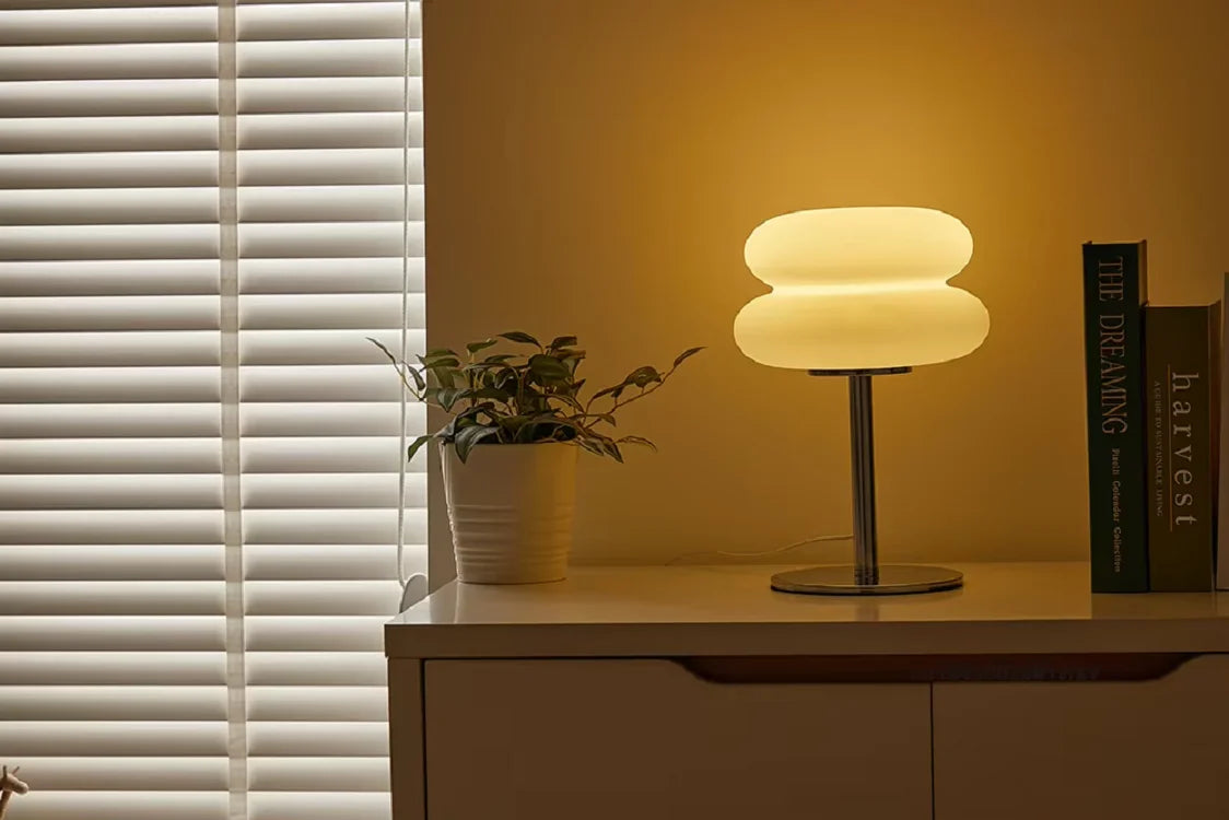 A modern cloud-shaped bedside table lamp with a light shade sits on a wooden surface next to a window blind.