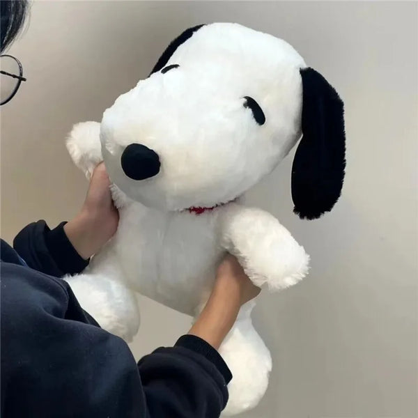 A person is holding a white Snoopy plush pillow with black ears and a black nose against a light background.