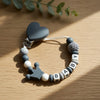 A silver clip featuring a heart-shaped silicone piece and a beaded bracelet with custom letters for a baby pacifier