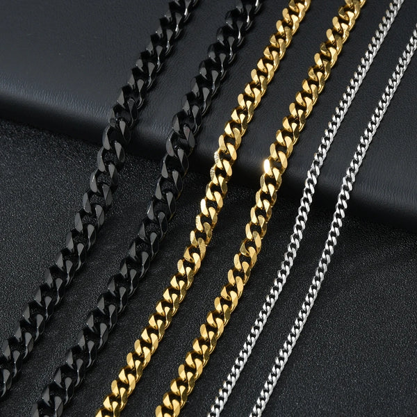 Trois colliers Cuban link en acier inoxydable noirs, or et argent empilés en diagonale sur une surface sombre.
