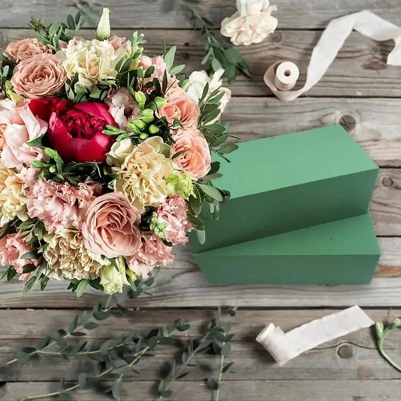 A square green box sits next to a lush arrangement of pink and peach roses and greenery on a wooden surface.