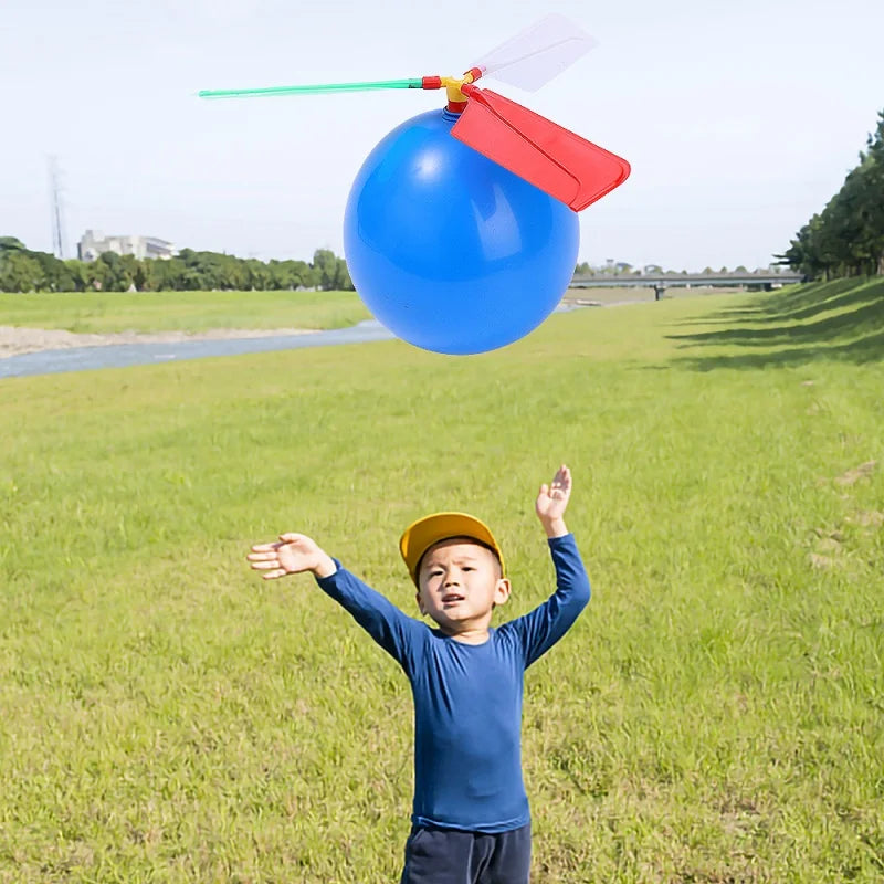 A young boy is throwing a blue propeller balloon helicopter toy into the air in a grassy field.