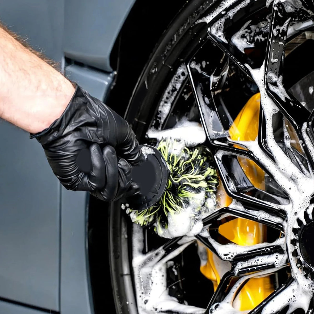 A hand wearing a black glove is cleaning a black wheel with a green brush inside a washing machine drum.
