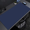 A dark blue oversized non-slip desk mat is placed on a gray desk next to a laptop and a small potted plant.