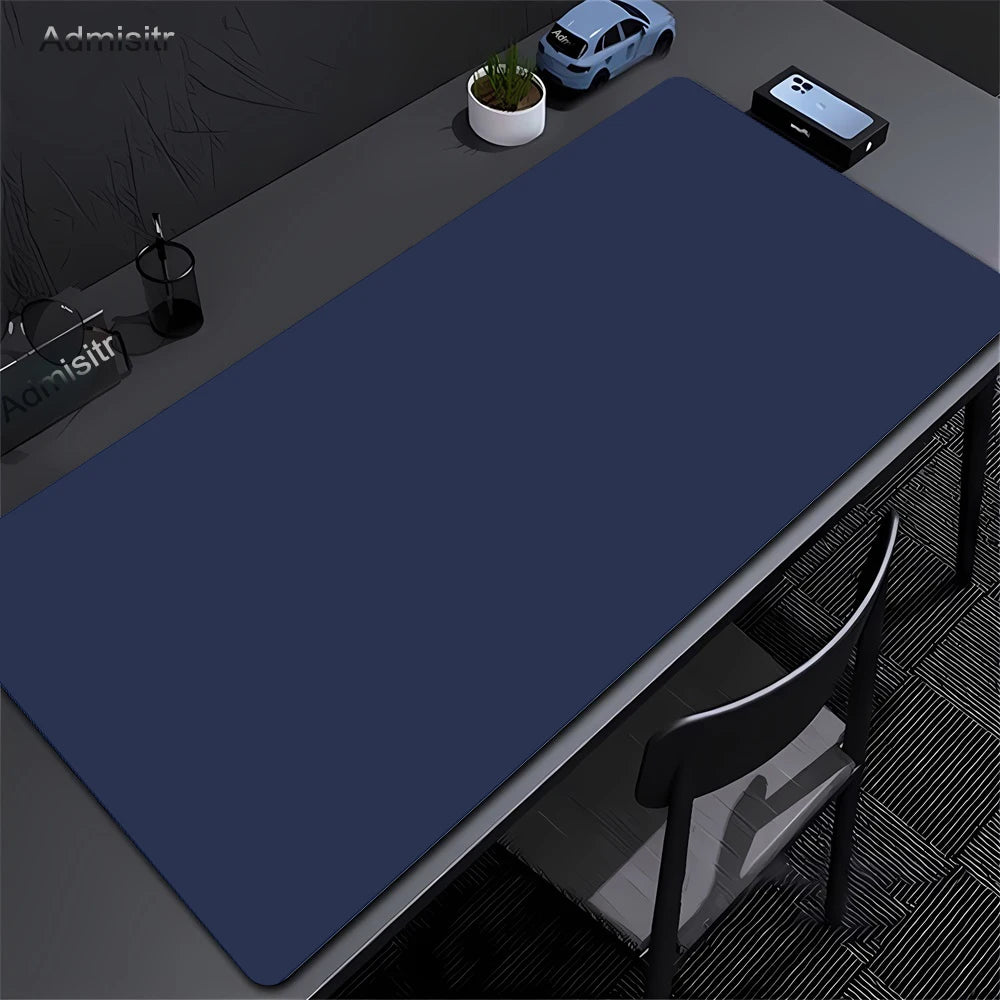A dark blue oversized non-slip desk mat is placed on a gray desk next to a laptop and a small potted plant.