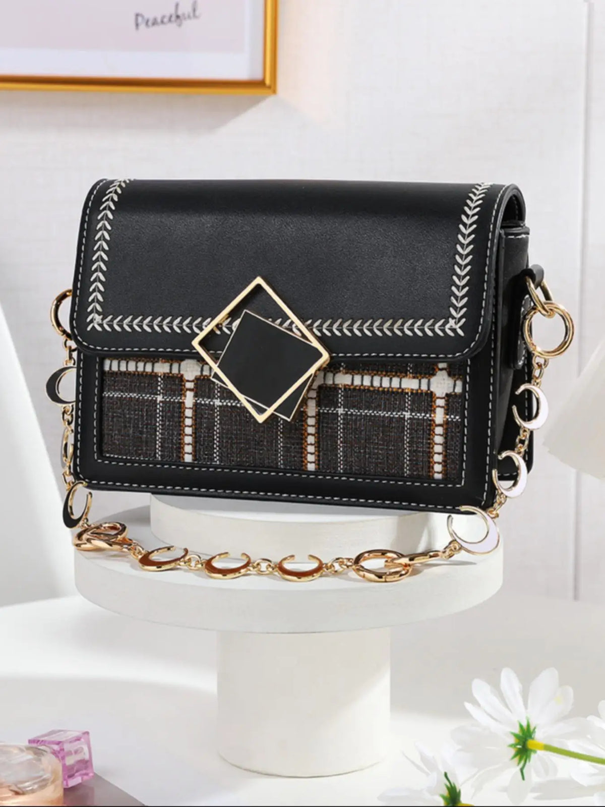 Schwarze Lederhandtasche mit goldenen Kettenverlängerungen und Zierstickerei auf weißem Ständer.