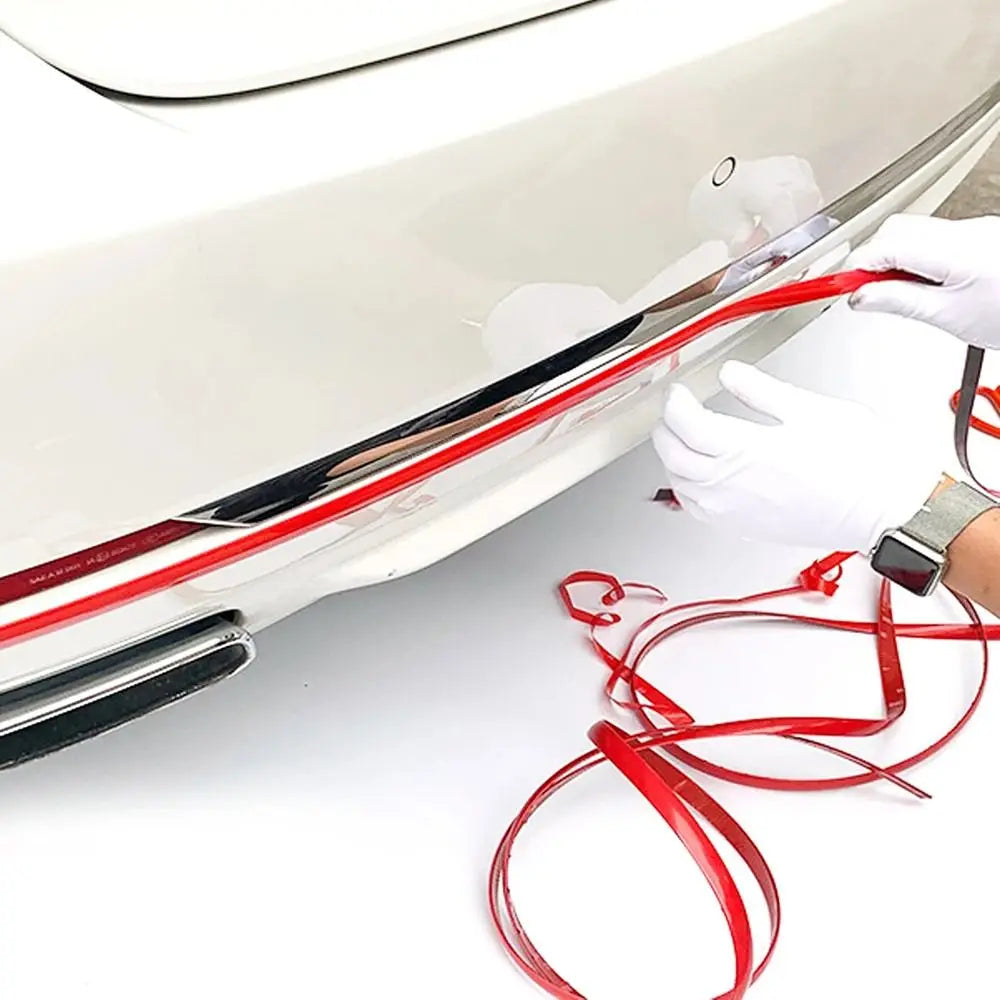 A hand is applying a flexible red PVC car bumper protection strip to a white car surface.