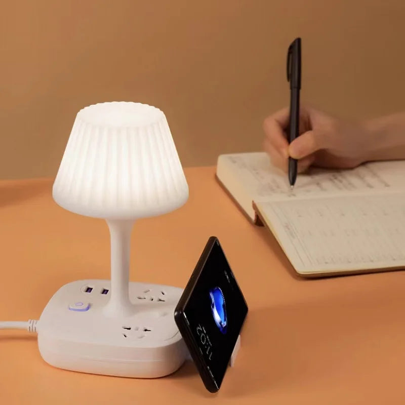 A white multifunctional desk lamp with a USB charging hub and phone holder is shown next to an open notebook and a pen.