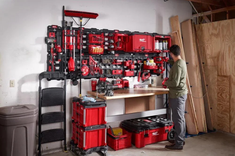 A red and black PACKOUT magnetic bin system mounted on a wall with tools and lumber in a workshop setting.