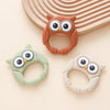 Three owl-shaped silicone teethers and rattles in orange, green, and white are displayed on a light background.