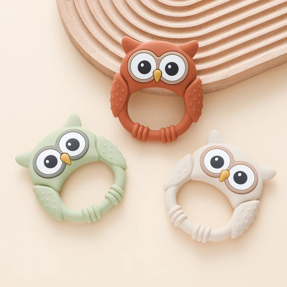 Three owl-shaped silicone teethers and rattles in orange, green, and white are displayed on a light background.