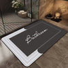 A rectangular dark gray bath mat with a white border and the word Bathroom printed on it, placed on a tiled floor.