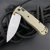 A light tan folding knife with a drop point blade is shown attached to a black cord on a dark surface.