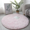 A fluffy pink shaggy circle area rug placed on a light gray floor next to a potted plant and a sofa.