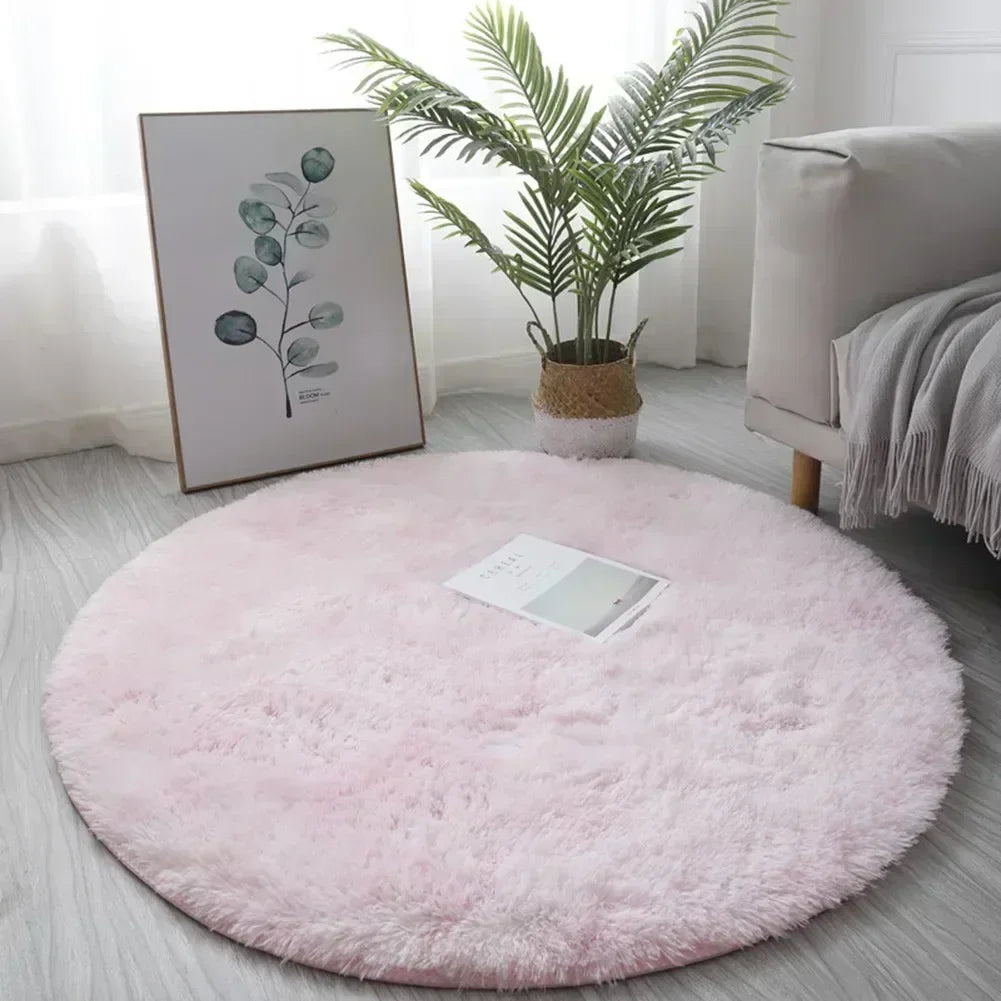 A fluffy pink shaggy circle area rug placed on a light gray floor next to a potted plant and a sofa.