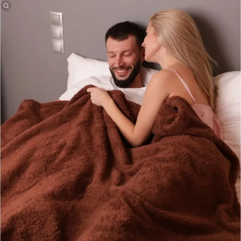 A smiling couple is wrapped in a large, fluffy brown throw blanket indoors.