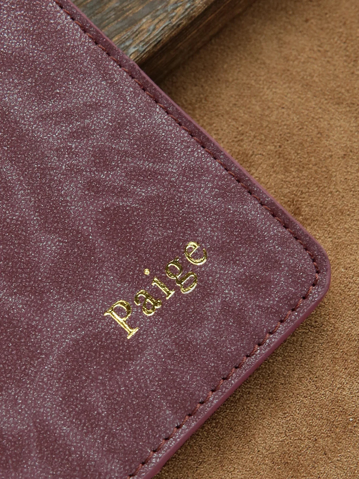 A deep plum colored leather passport holder with gold embossed name on a textured brown background.