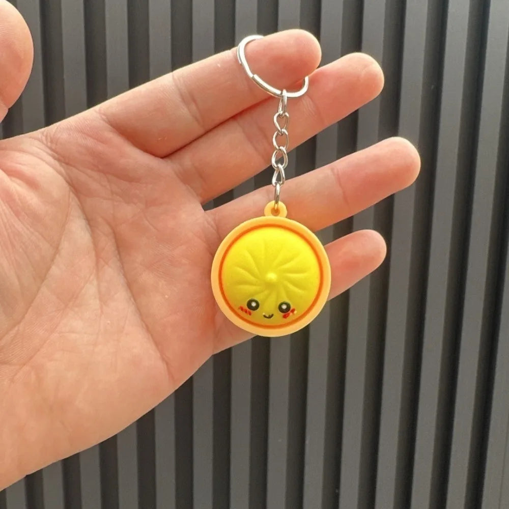 Eine Hand hält einen gelben, lächelnden, dumplingförmigen Schlüsselanhänger mit einem silbernen Ring.