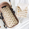 A beige and brown cozy cartoon bear stroller cushion is shown next to a stack of cream-colored fabric items.
