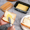 A hand holds a slice of bread with a stainless steel knife, next to a black metal butter dish labeled Butter.