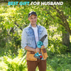 A man wearing a baseball cap and denim shirt holds a blue cordless mini chainsaw in an outdoor garden setting.