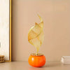 A golden, abstract sculpture shaped like a leaf stands on an orange ceramic sphere.