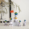 A collection of handcrafted astronaut figurines in white and silver on a white pedestal with a small orange flag.