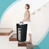 A woman stands next to a large black laundry basket on a wooden shelf, holding a blue item.