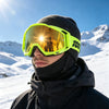 A person wearing bright yellow-green off-road motorcycle goggles and a black beanie is standing in a snowy mountain landscape