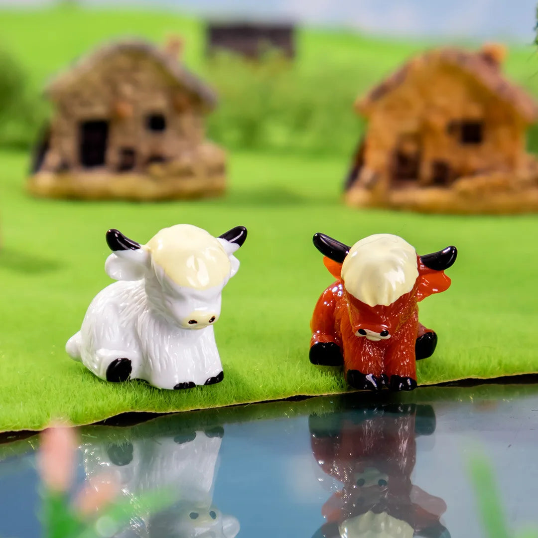 Two adorable resin Highland cow figurines, one white and one orange, displayed on green grass.