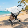 A beige and white stretchable UV-protective stroller sun shade is shown on a paved path near a tropical beach.