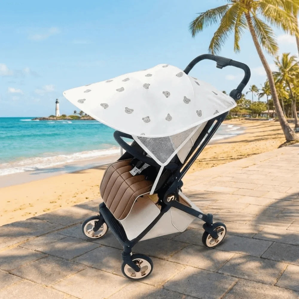 A beige and white stretchable UV-protective stroller sun shade is shown on a paved path near a tropical beach.