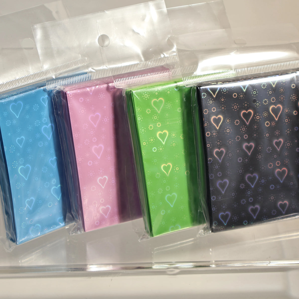 A collection of holographic heart-shaped card sleeves in various pastel and bright colors displayed in clear plastic packagin