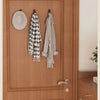 A wooden door features three hooks displaying a black and white patterned towel, a gray robe, and a round woven mat.